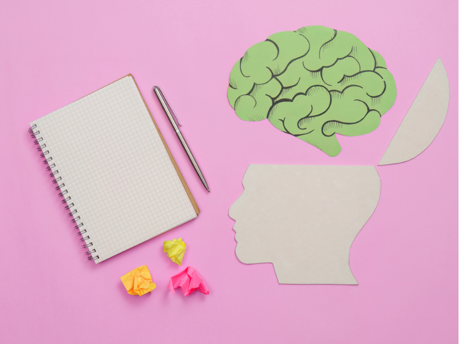 Recorte de una cabeza infantil con un cerebro de papel verde separado, junto a una libreta, un bolígrafo y papeles de colores sobre fondo rosa.